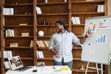 Young African American employee standing explaining project results in graphs, streaming lesson online and looking at webcam, pointing on whiteboard, talking with the virtual group online via laptop