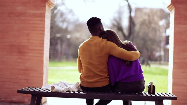 Friend embracing girlfriend, black man putting arm around girl sitting at park bench
