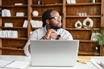 Serious thoughtful young African American man with dreadlocks sitting at desk in the office library and looking away, taking break from working on laptop, pondering project plan, thinking of new ideas
