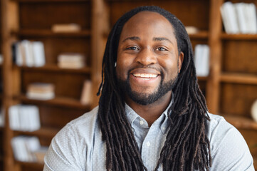 Portrait close-up of a smiling young handsome African American man with dreadlocks standing posing in the library or and looking at the camera, the optimistic black man office manager is ready to work