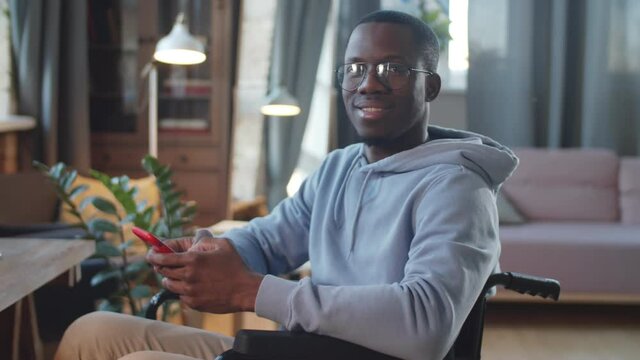 Waist up shot of young disabled Afro-American man on wheelchair holding smartphone, looking at camera and smiling in living room at home