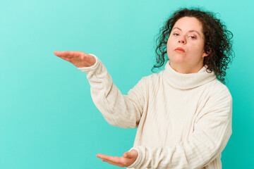 Woman with Down syndrome isolated shocked and amazed holding a copy space between hands.