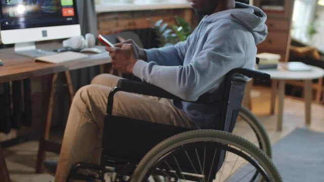 Zoom in shot of paraplegic Afro-American man on wheelchair messaging on smartphone while working remotely from home