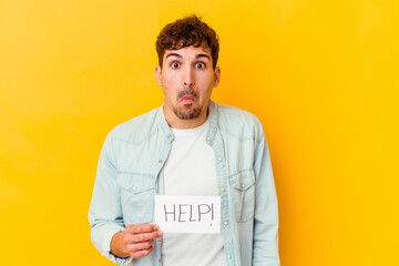 Young caucasian man holding a help placard isolated shrugs shoulders and open eyes confused.