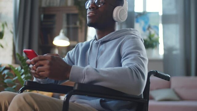 Tilt up shot of young Afro-American man on wheelchair listening to music on smartphone with wireless headphones and smiling at home