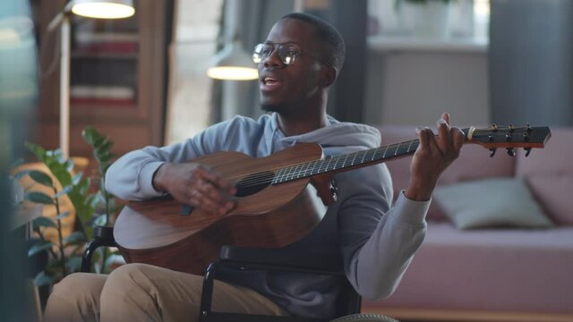 Young paraplegic Afro-American man on wheelchair playing the guitar and singing a song at home