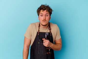 Young barber caucasian man isolated on blue background shrugs shoulders and open eyes confused.
