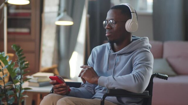 Young disabled Afro-American man on wheelchair listening to music on smartphone with wireless headphones, looking at camera and smiling at home