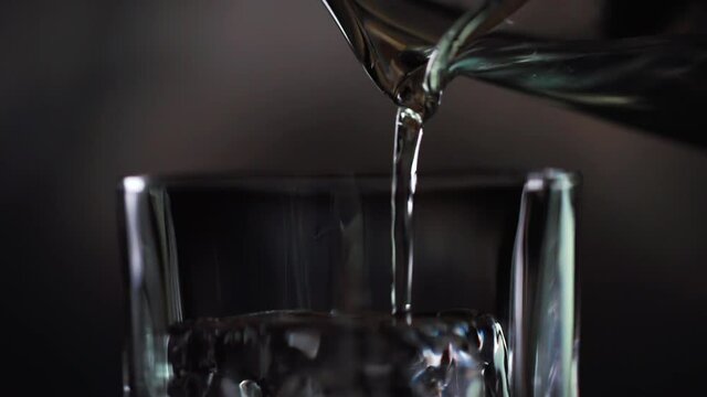 Water or lemonade is poured into glasses with ice on a macro plan from the spout of another glass. On a black background