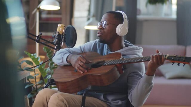 Young Afro-American man on wheelchair in wireless headphones playing the guitar and singing in microphone while recording song in home studio
