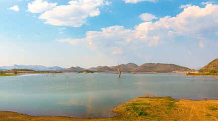 Massive reservoir and surrounding dry mountainous landscape with white clouds and blue sky reflected in the still waters created by the dam.
