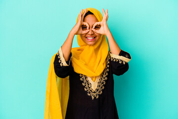 Young muslim woman isolated on blue background showing okay sign over eyes