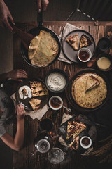 A woman holds a frying pan with freshly baked thin pancakes, a child eats pancakes, on a festive table pancakes and sauces to them On a dark wooden background.