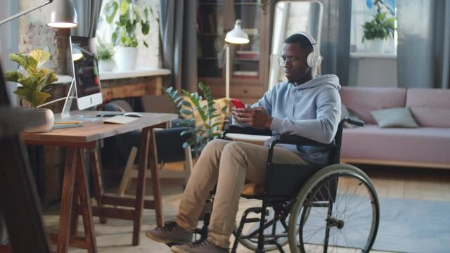 Young paraplegic Afro-American man on wheelchair using smartphone and listening to music with wireless headphones at home