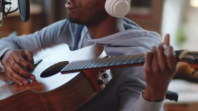 Tilt up shot of young disabled black man on wheelchair in wireless headphones singing into microphone and playing the guitar while recording music at home