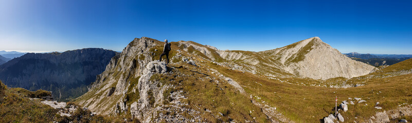 Fototapeta premium A man with a hiking backpack standing at the edge of a high mountain, Hohe Weichsel in Austria. The man in enjoying the calmness and peace. Lush green pasture around him. Exploration and discovery
