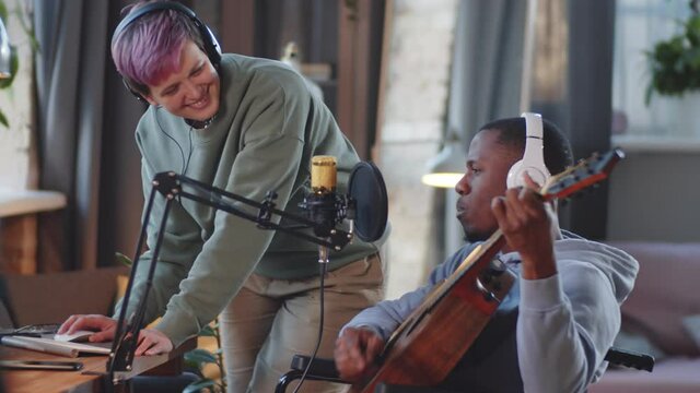 Cheerful Afro-American man on wheelchair playing the guitar and singing in microphone while recording song with help of joyous female friend in home studio