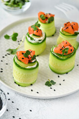 Cucumbers rolls with soft cheese, pieces of salted salmon, microgreens and black sesame served on a white plate. Holiday vegetable appetizers. Selective focus