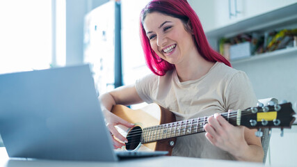 Fototapeta na wymiar A woman sits in the kitchen during a remote acoustic guitar lesson. A girl learns to play the guitar and watches educational videos on a laptop