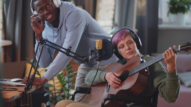 Young disabled woman on wheelchair playing the guitar and singing in microphone while recording song with assistance of cheerful Afro-American male friend in home studio