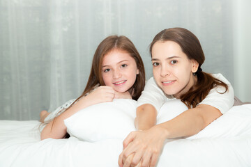 Portrait happy mother and teen daughter smiling and looking at the camera, lying on bed together....
