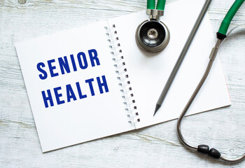 SENIOR HEALTH is written in a notebook on a wooden table next to stethoscope.