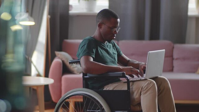 Young disabled Afro-American man on wheelchair typing on laptop in living room while browsing the Internet at home