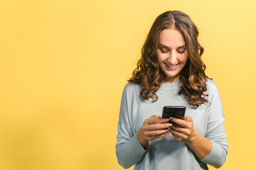 Cheerful attractive brunette woman holding smartphone, enjoying chatting in social networks, texting messages and smiles, using mobile app for dating on the phone, isolated on yellow, copy space