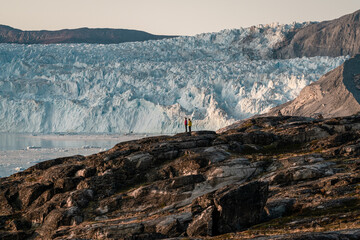 Unidentified People sitting standing in front of huge glacier wall of ice. Eqip Sermia Glacier Eqi...