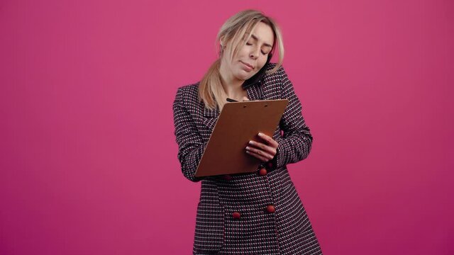 Businesswoman, attentive writes in the notebook an important information and speaks on the phone, is focused. Beautiful young mature blonde in pink jacket. Isolated on a pink background. Concept of
