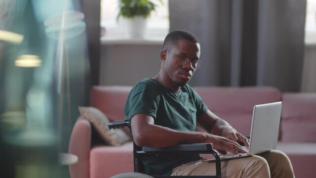 Cheerful disabled Afro-American man on wheelchair using laptop in living room at home and then looking at camera and smiling