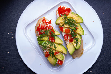 two canapes, slices of fresh avocado and slices of ripe tomato are laid out on a slice of baguette and sprinkled with sesame seeds, on a white plate