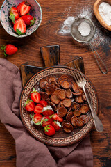 Mini chocolate pancake cereal with strawberries for breakfast on old wooden table. Trendy home breakfast with tiny pancakes. Top view