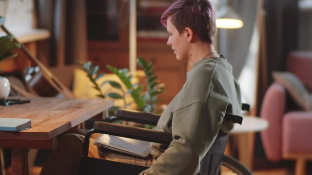 Side view of young disabled woman riding wheelchair to desk, opening laptop and typing on it while working online at home