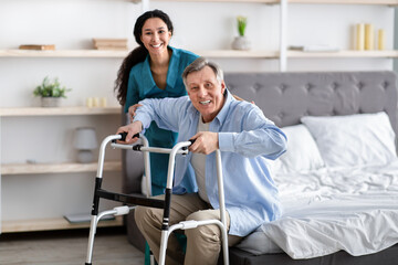 Female nurse helping elderly male with walking frame stand up from bed at home. Professional care...