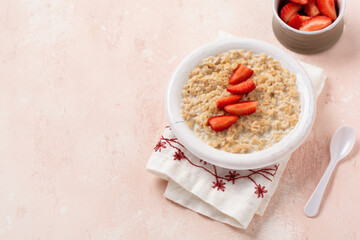 Simple oatmeal porridge with strawberries in a white plate on a linen napkin on pink background. Breakfast health food concept. Top view