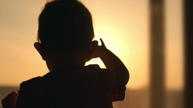Anxious child knocks his hand on the window while standing inside the house during sunset