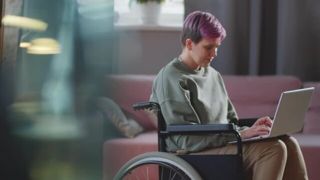 Positive disabled woman on wheelchair typing on laptop at home and then looking at camera and smiling
