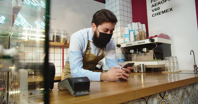 Portrait Of Good-looking Male Small Business Owner In Medical Protective Mask. Young Caucasian Man Using Smartphone To Take Orders, Typing And Looking Through Social Medias. Technology Concept.