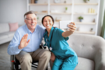 Fototapeta premium Cheerful disabled senior man in wheelchair and his nurse taking selfie indoors, selective focus