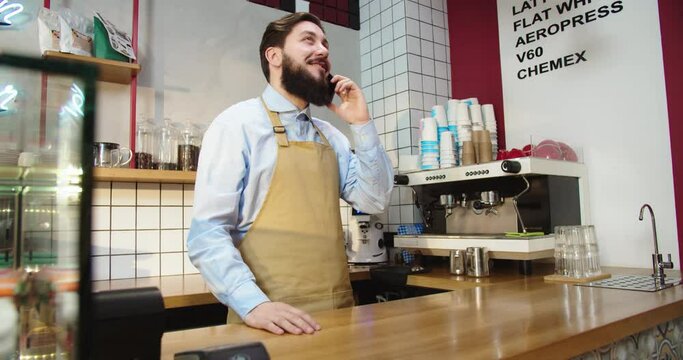Attractive Caucasian man talking on phone to customer and taking pre-orders. Young male barista working adn consulting clients on beverages menu in coffeehouse. Business, communication concept.