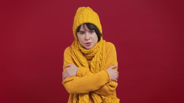 Sad sick ill short hairdo young woman 20s years old in yellow knitted sweater hat scarf looking camera rubs her hands frozen chills shivers cold season isolated on bright vivid red background studio