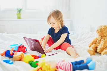 Toddler girl with laptop, notebook, phone and different toys in bed on clean white linens