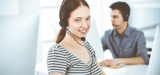Fototapeta na wymiar Casual dressed young woman using headset and computer while talking with customers online. Group of operators at work. Call center, business concept