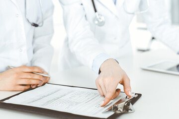 Unknown woman-doctors at work with patient at the background. Female physicians filling up medical documents or prescription while standing in hospital reception desk, close-up. Health care concept