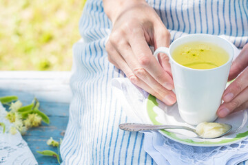 Woman hands keep white cup with fresh linden flowers tea and plate with spoon and honey on a stripped apron background outdoors, person rests on a blue colored bench in the garden