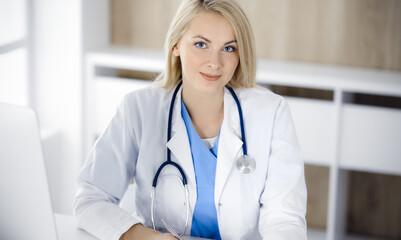 Portrate of woman-doctor at work while sitting at the desk in clinic. Blonde cheerful physician ready to help patients