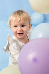 Fototapeta na wymiar Beautiful cheerful little girl 1 year old with blue eyes in white turtleneck among balloons and looks at the camera