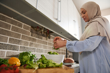 Woman in hijab tears lettuce leaves with her hands and throws them into a bowl of vegetables to make a salad