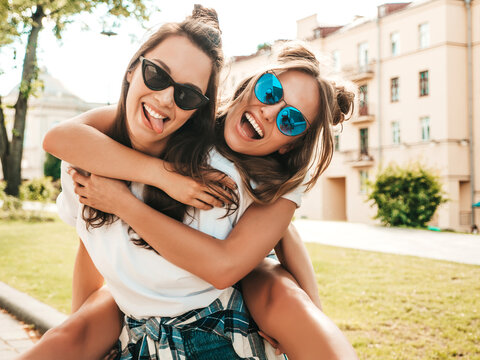 Two young beautiful smiling hipster female in trendy summer white t-shirt clothes.Sexy carefree women posing on street background. Model jumping on her friend back, gives piggyback riding outdoors